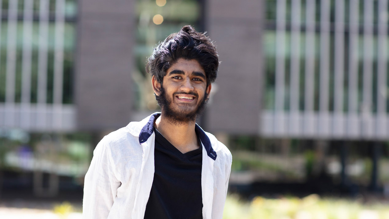 Ali has dark skin, black hair and a black beard and moustache. He is standing on UOW campus, wearing a black t-shirt and white overshirt.