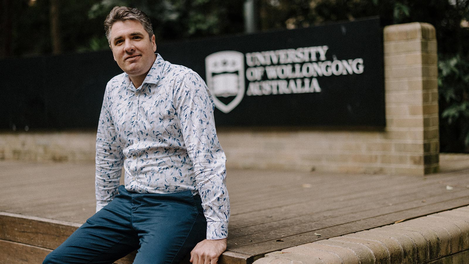 Professor Andrew Ainsworth has short, grey hair and is sitting on a brick stage in front of a black and white University of Wollongong sign. He is wearing a white floral dress shirt and blue dress pants.