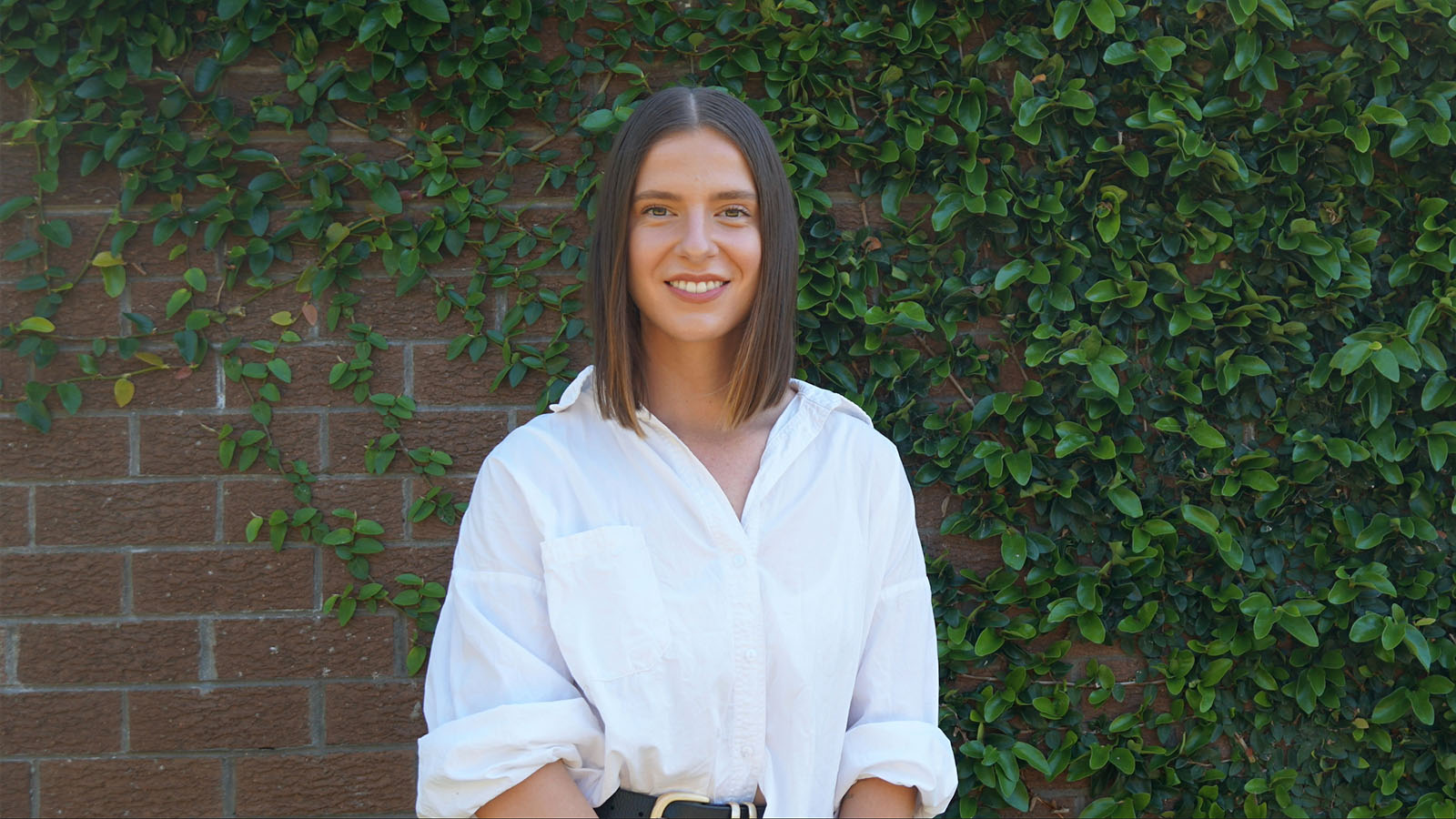 A woman in a white button-up shirt it standing in front of a brick wall covered in vines, smiling.