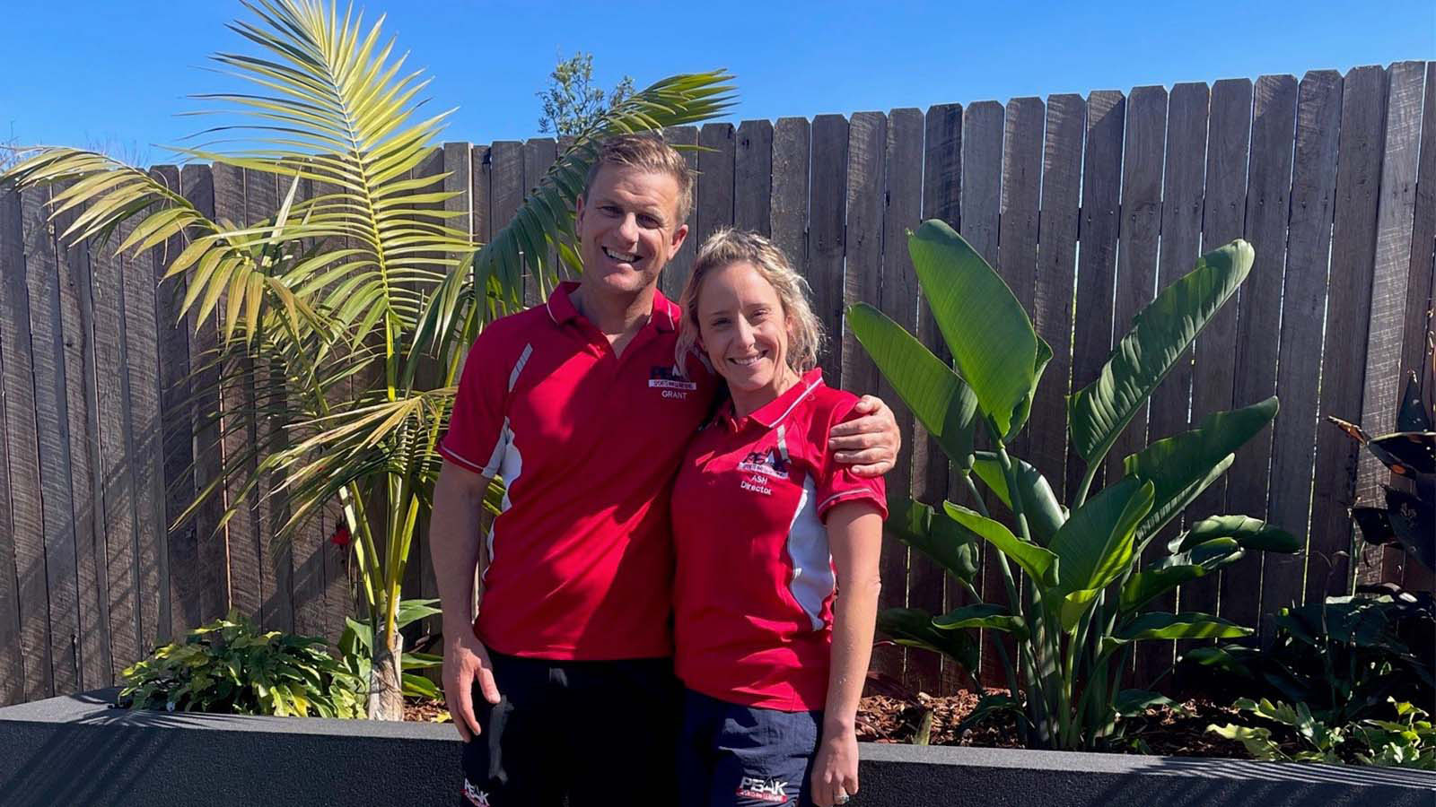 Grant and Ashleigh Neill are standing in a backyard wearing red polo shirts.