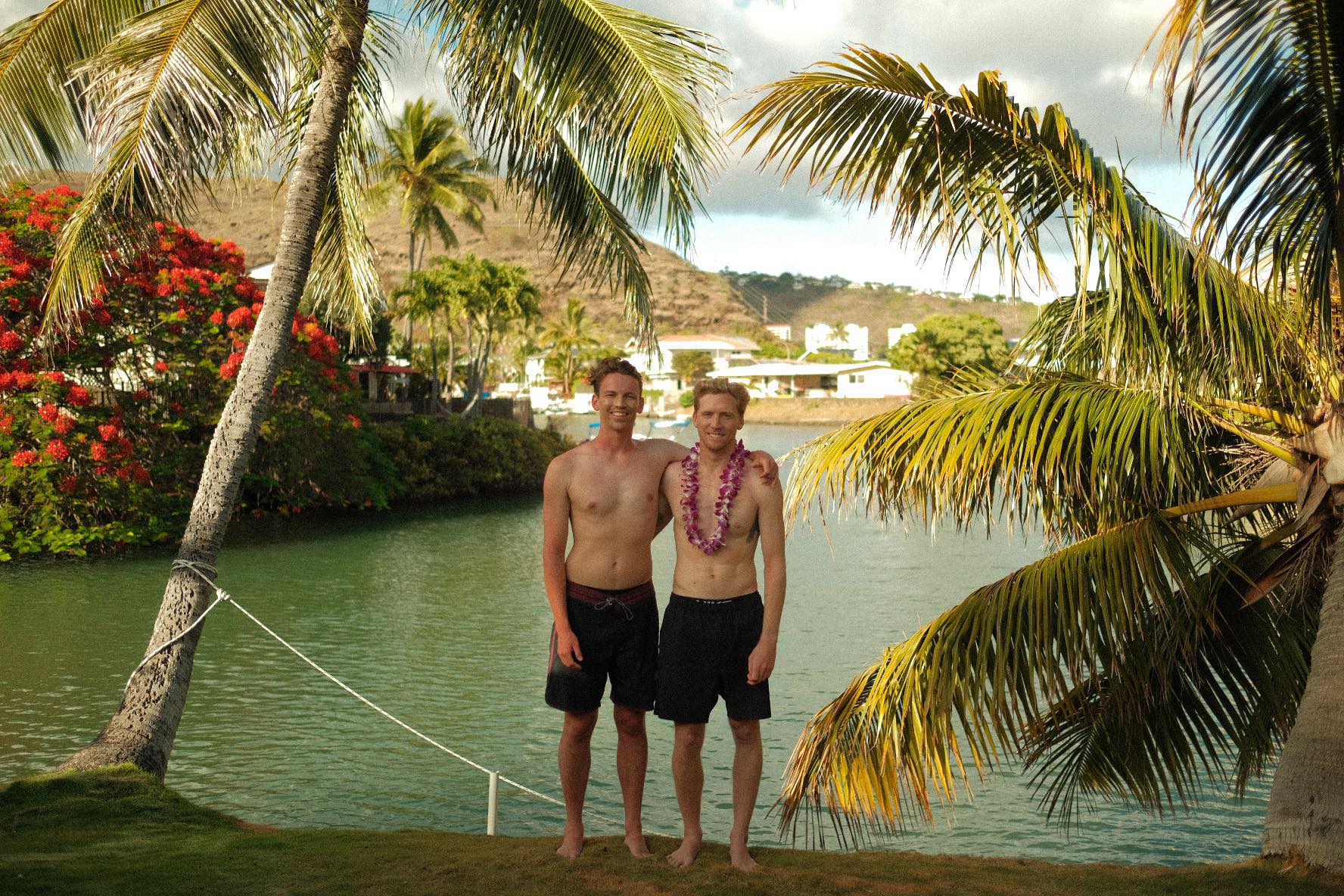 Sam Tolhurst and Brett Connellan are standing on an beach. They have their arms around each others shoulders smilinng, and Brett is wearing a lei around his neck.
