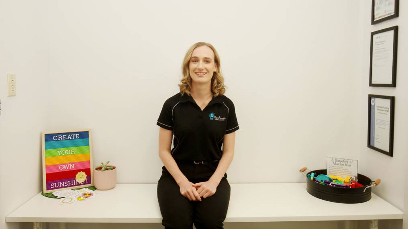 A young woman is sitting on a table in a classroom. Awards are hung on the wall to her left, and there is a rainbow sine that reads 'Create your own sunshine' on her right.