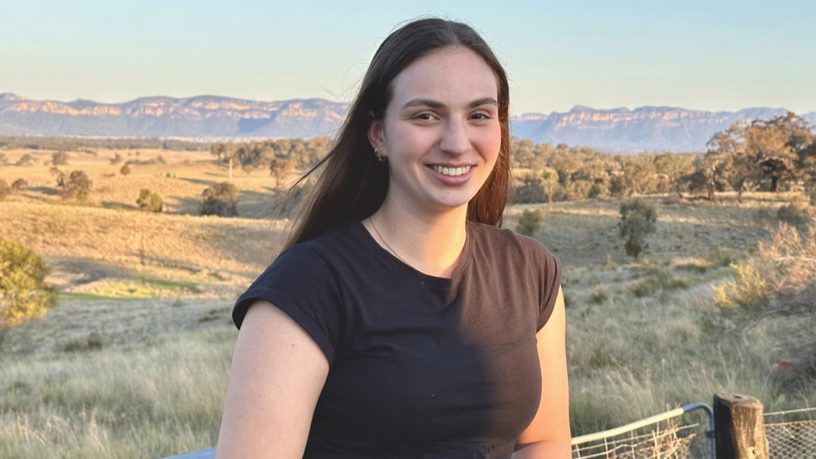 A young woman is standing on a rural property, smiling.