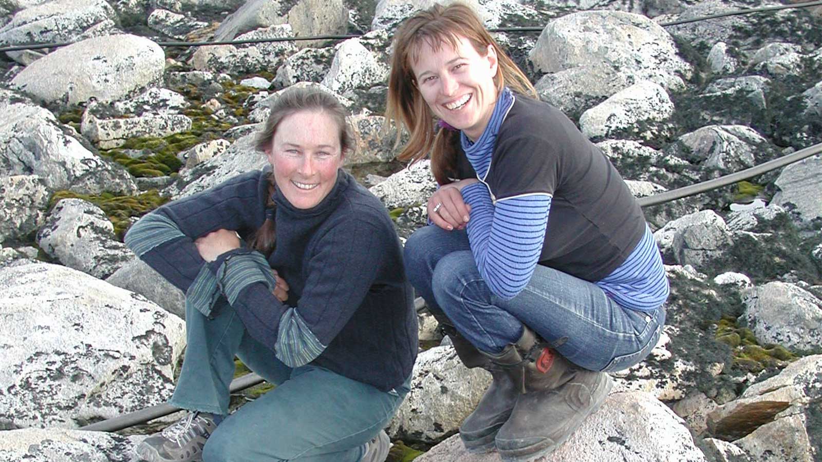 Two women are sitting on rocks in Antarctica. They are wearing long sleeve jumpers, blue jeans and hiking boots. They are squatting down smiling.