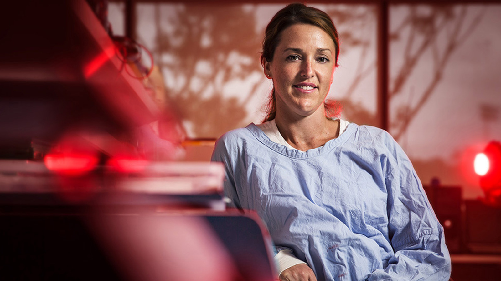 Dr Katrina Green is in a lab, wearing a blue lab gown, seated at a bench leaning against it and smiling.