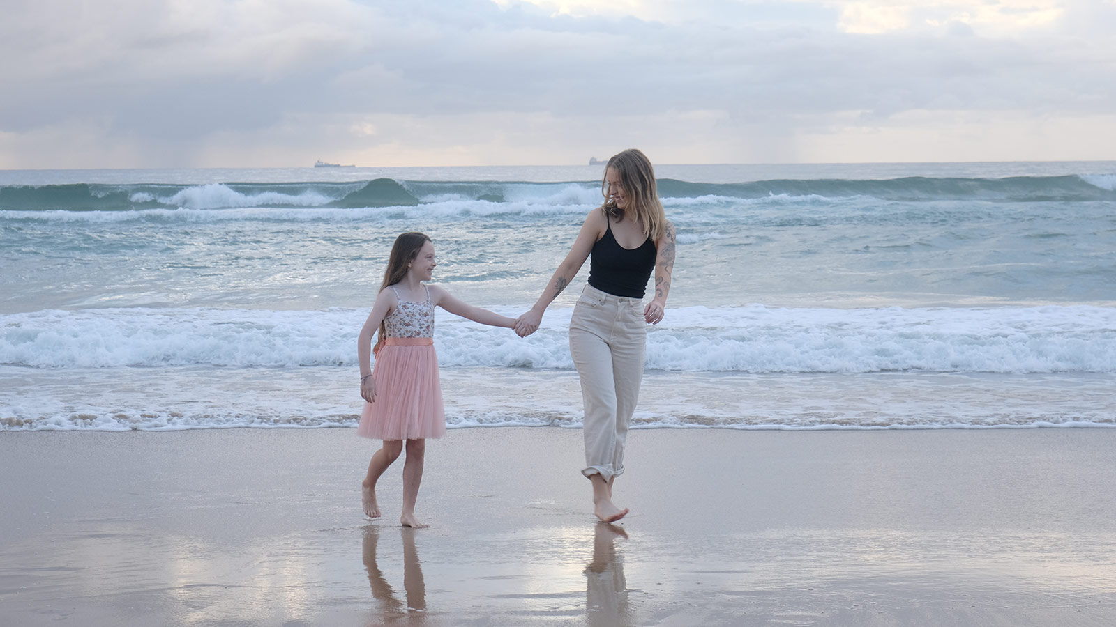 Lara Warwick and her daughter are holding hands on a beach at dusk. They are standing in the shore smiling at each other. Lara is wearing a black top and white pants, her daughter is on her left wearing a pink floral dress.