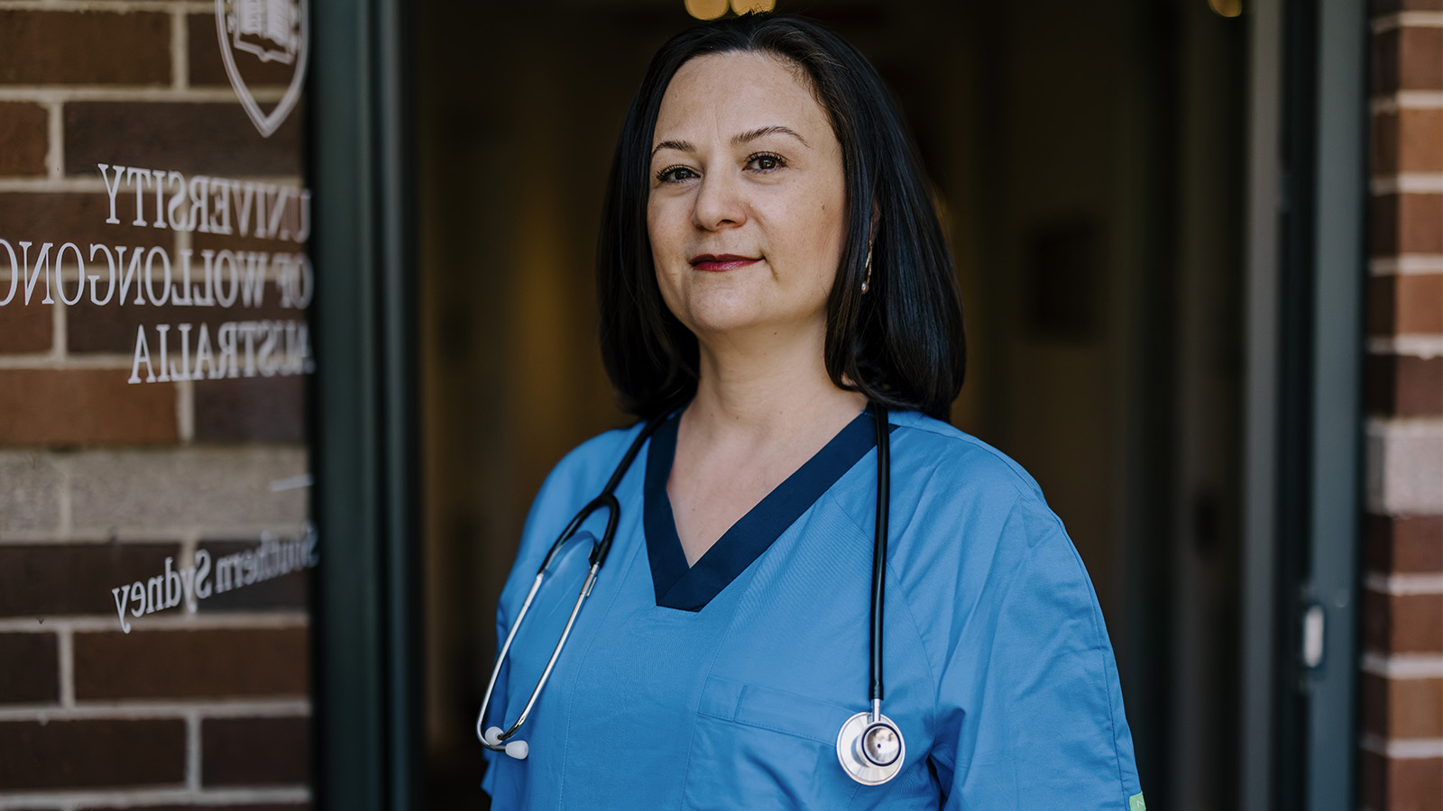 Laureine Gabriel is standing in front of the UOW Sutherland door wearing blue scrubs and a stethoscope.