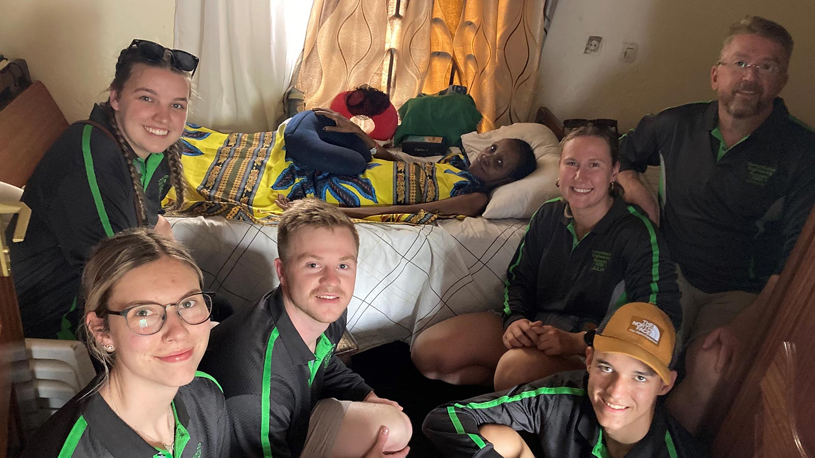 A group of people in black and green shirts are sitting around woman laying in bed.