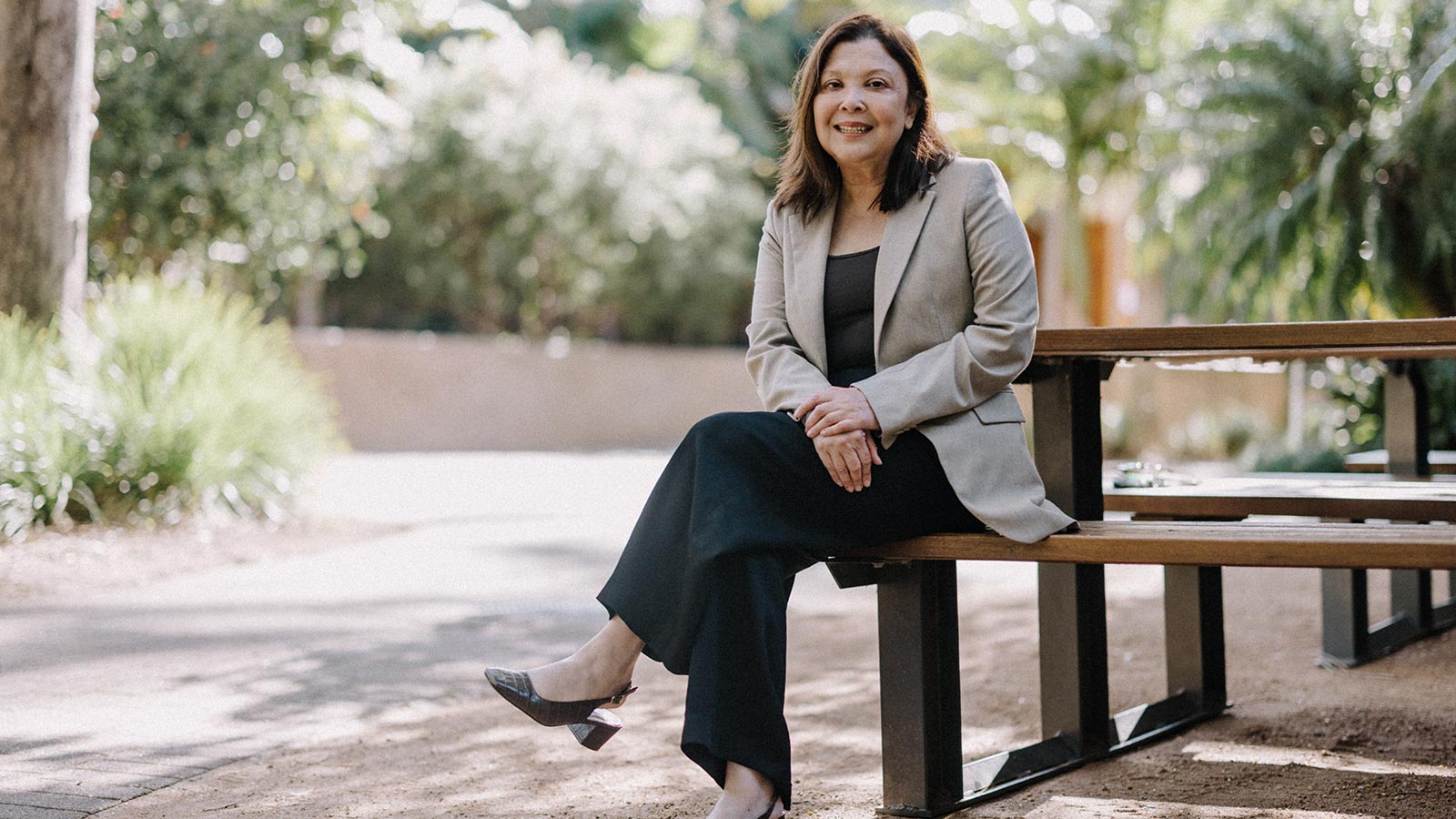 Professor Millicent Chang is wearing a black top, pants and shoes with a light grey dress jacket. She is sitting on a bench on UOW with her legs crossed, smiling.