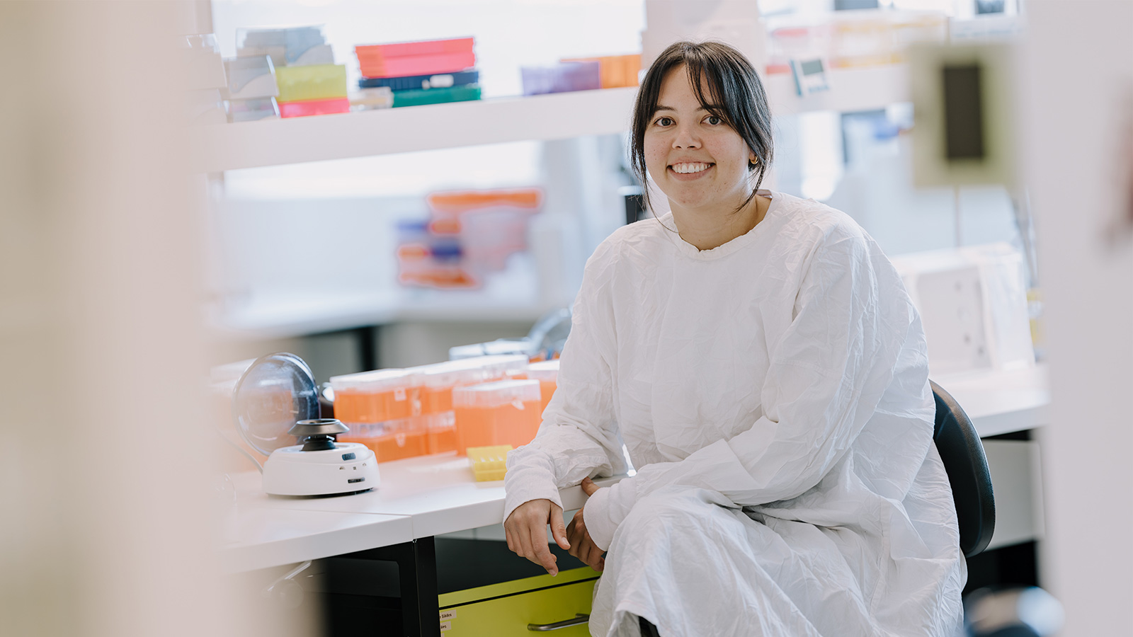 A woman with dark hair is sitting at a lab bench, wearing a lab coat.