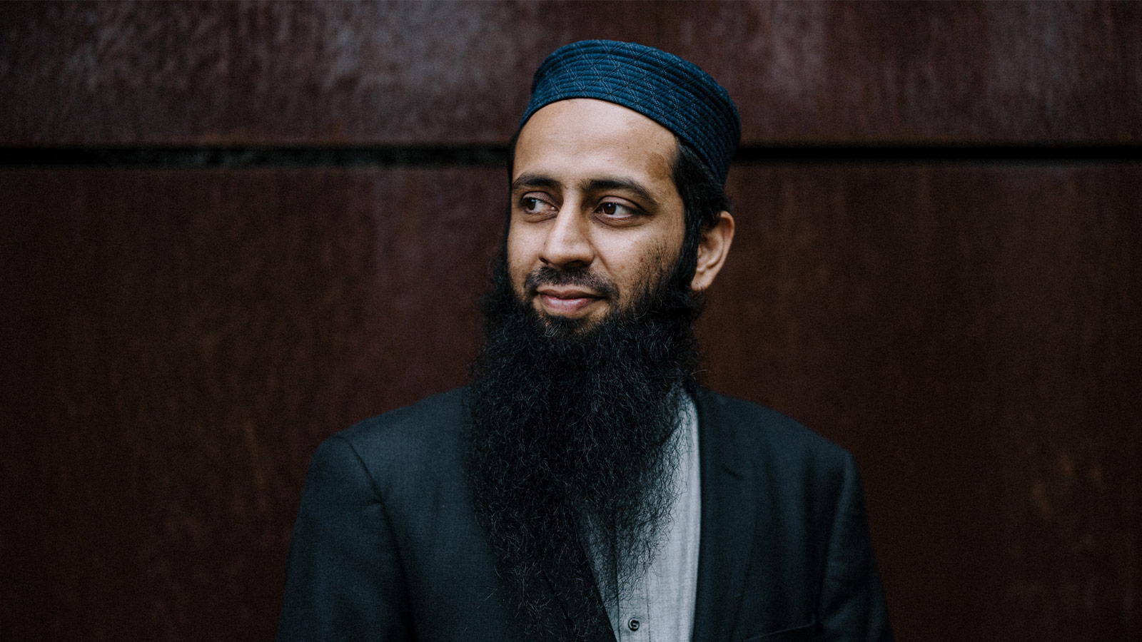 Professor Searat Ali has a long, dark beard. He is wearing a light blue shirt and dark jacket, and a dark prayer cap. He is standing against a dark brown metal wall.