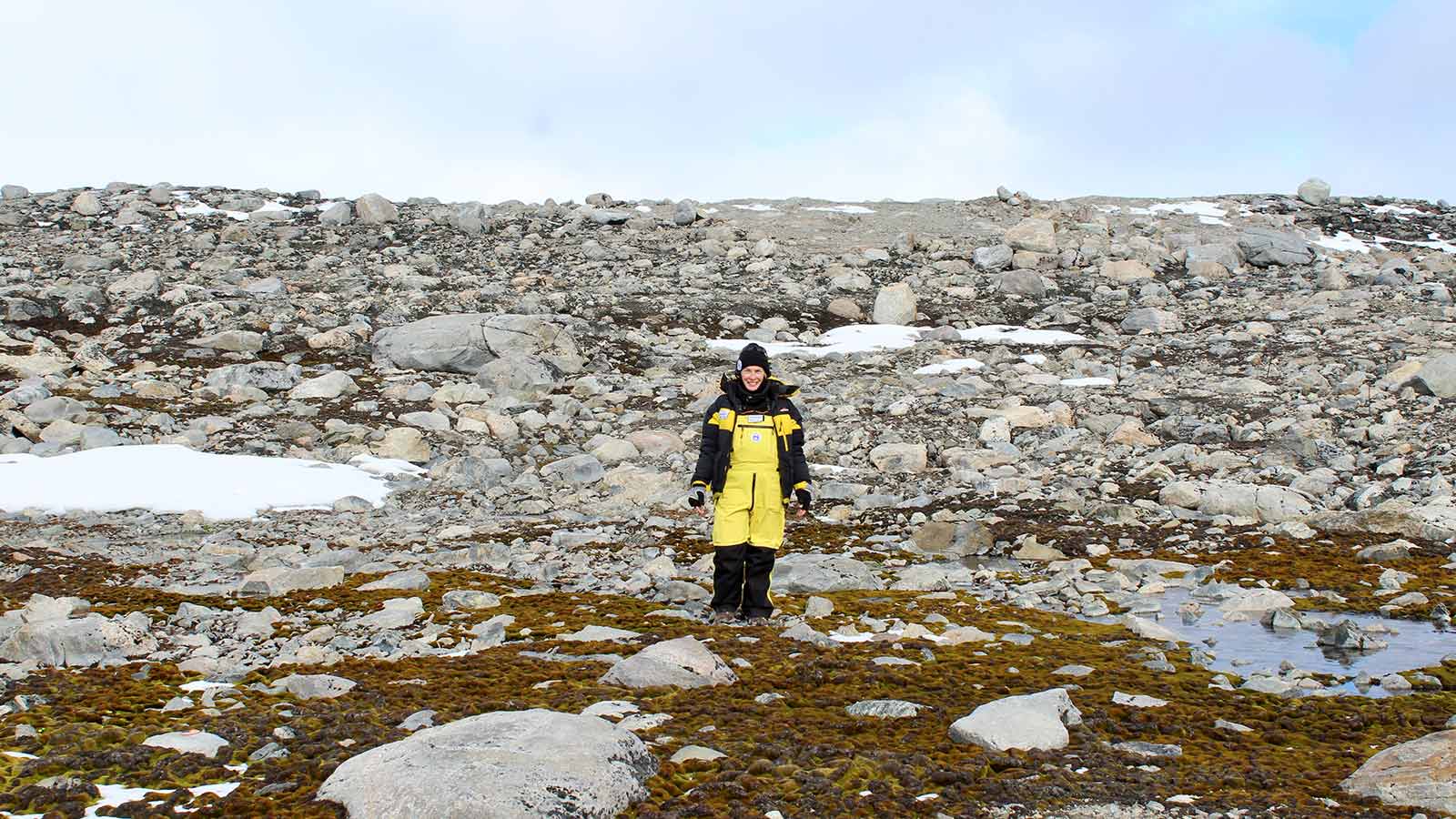 Professor Sharon Robinson is standing in front of moss, rocks and ice in Antarctica. She is wearing black and yellow protective thermals, beanie and gloves and smiling.