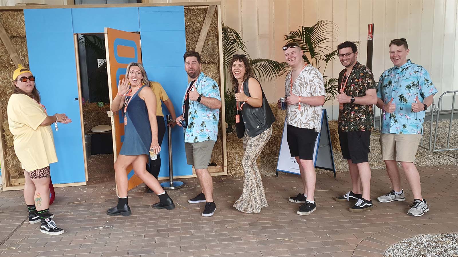 A line of festival patrons are lining up for a compost toilet made out of wood and hay bales. They are smiling at the camera holding their thumbs up.