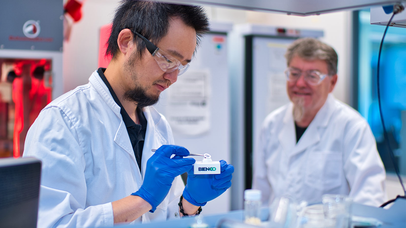 Two men in a science lab. They are both wearing lab coats and safety goggles. The man in the foreground is wearing blue latext gloves, holding tweezers to place a 3D printed film in place while the man in the background supervises.