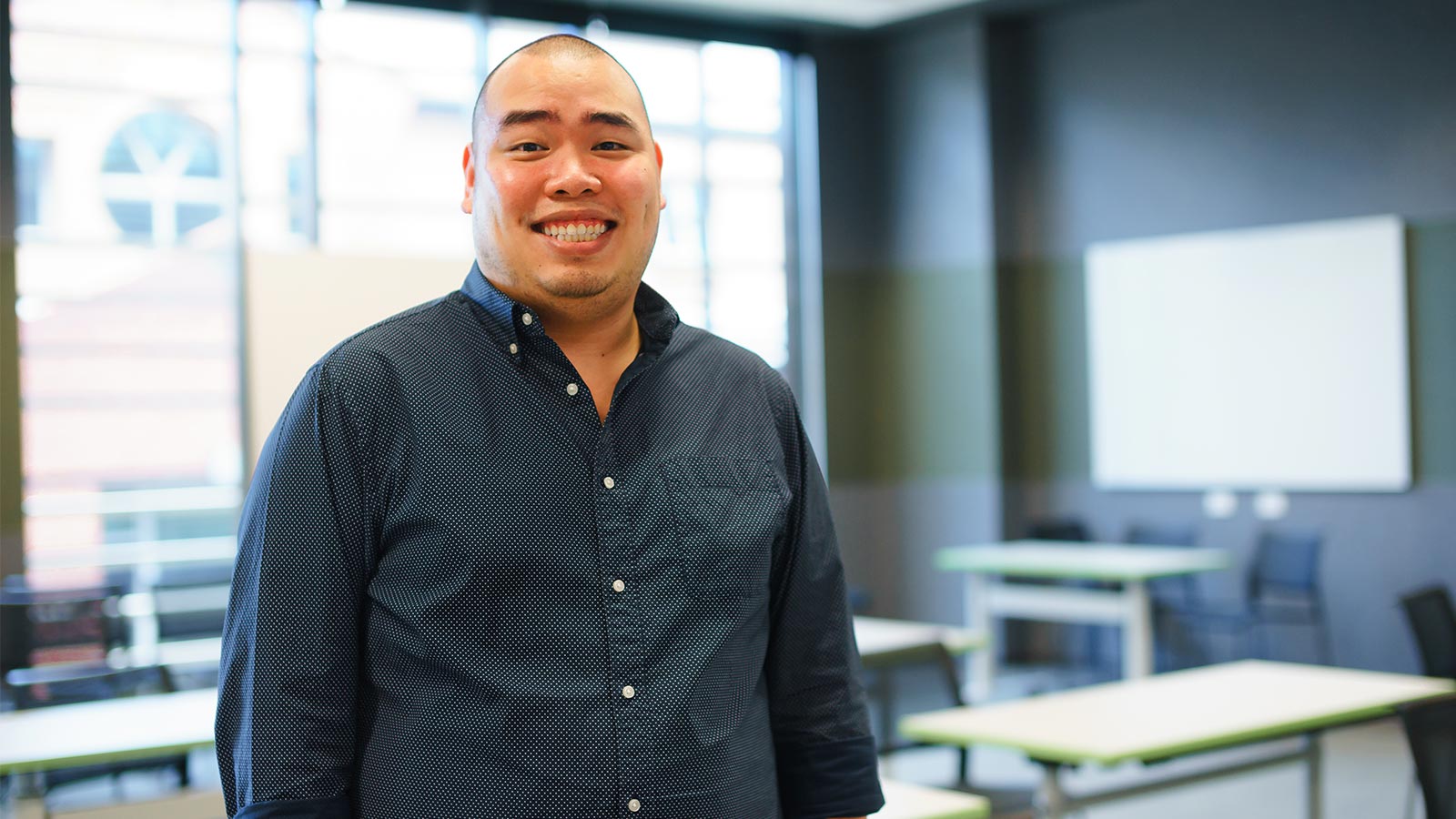 Dr Beo Thai is standing in a classroom at university. He is wearing a dark blue, collared, button-up shirt and smiling.