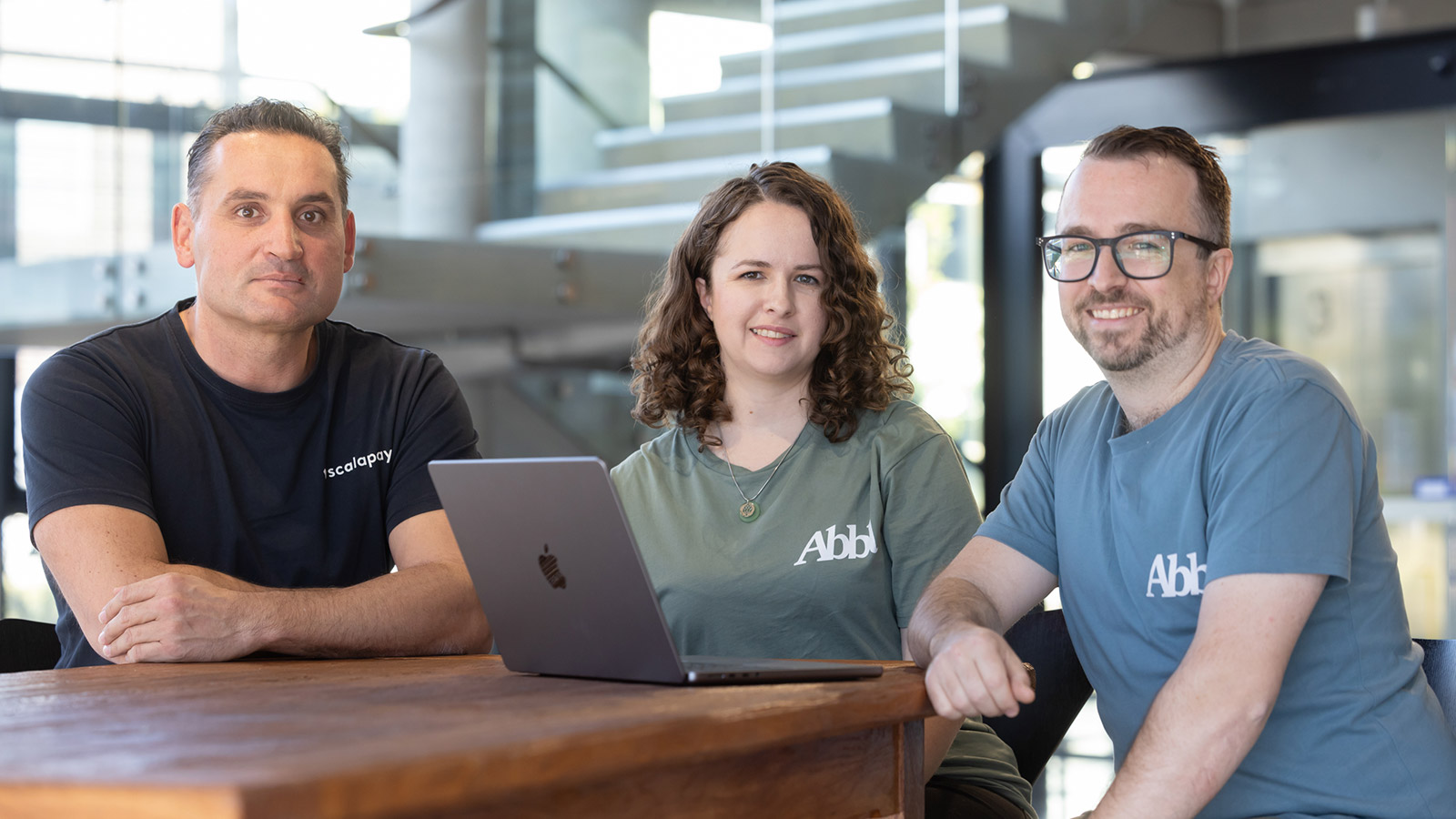 Two men and a woman are sitting at a wooden bench table with a laptop