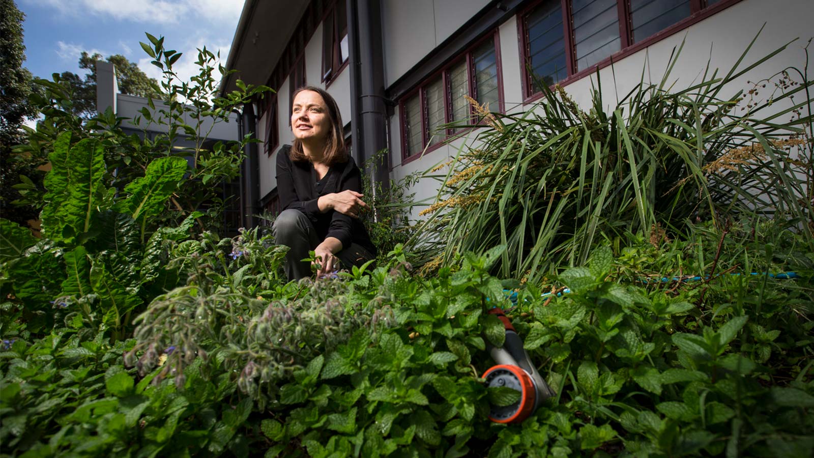 Natascha has brown shoulder length hair and is wearing a black top and jeans. She is crouched down in a garden plucking a flower.