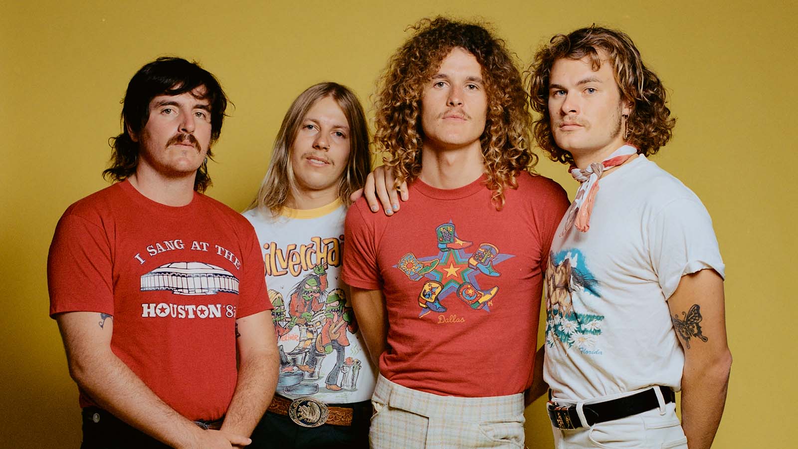 Four young men in t-shirts and pants against a yellow backdrop.