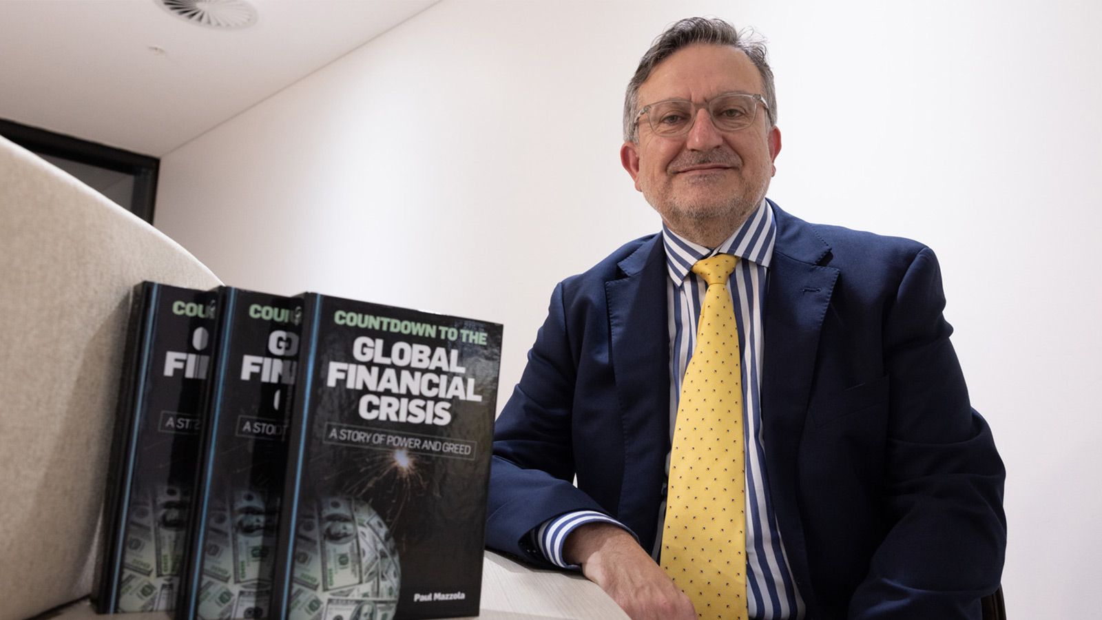 Paul Mazzola is Paul Mazzola is wearing a navy blue suit and yellow tie. He is sitting at a desk next to standing copies of a book titled ‘Countdown to the Global Financial Crisis’.wearing