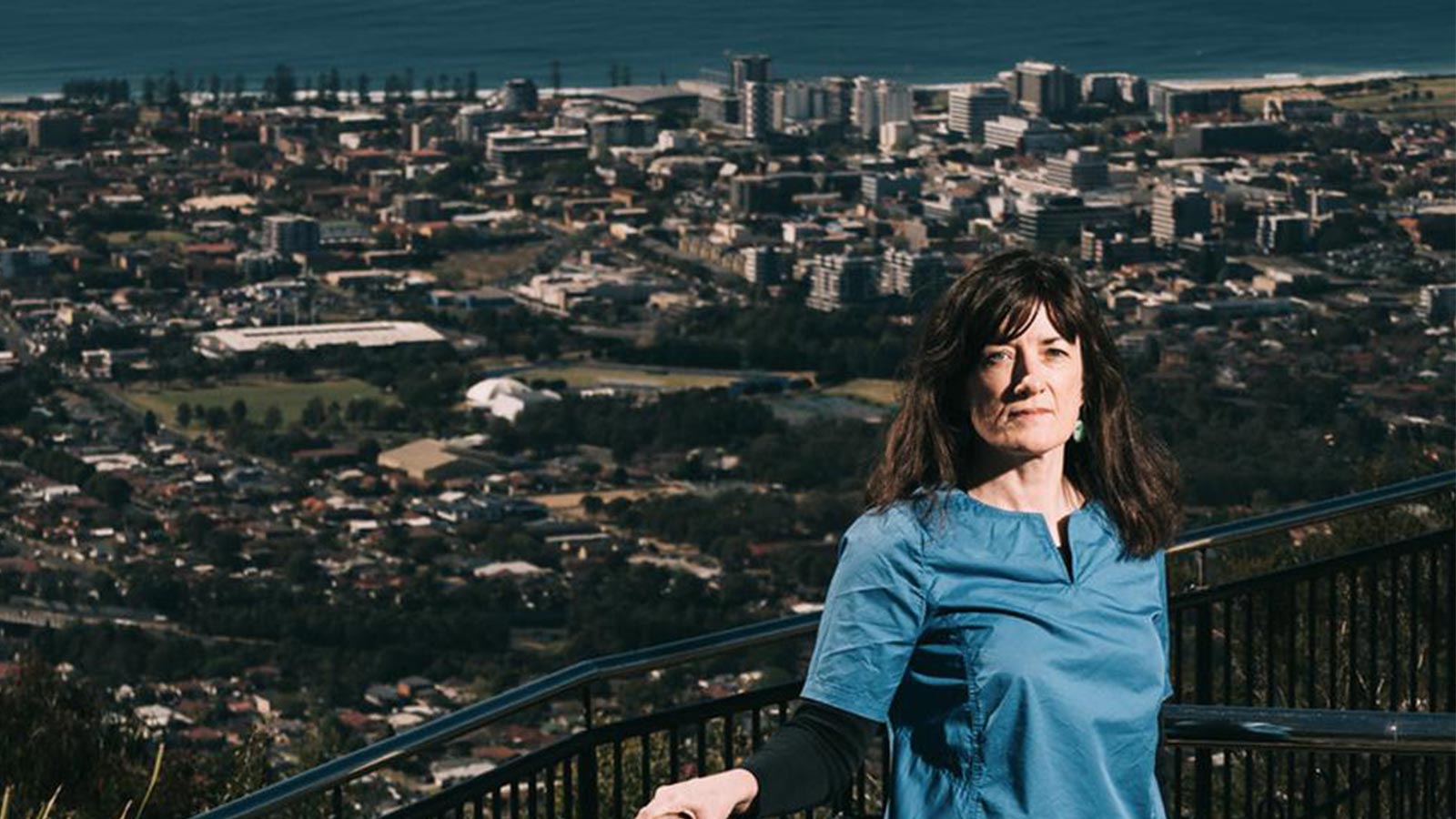 Pauline McGuirk has dark shoulder length hair and a blue T-shirt. She is leaning against a fence at Mount Keira, which overlooks the Wollongong housing and beach skyline.