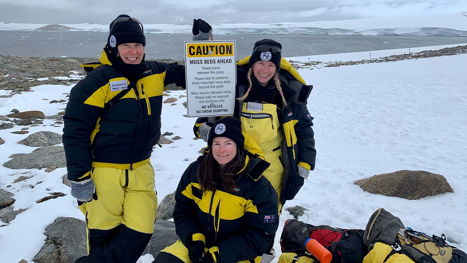 Three women are standing in the snow of Antarctica. They are each wearing black and yellow protective thermals, beanies and gloves. One is leaning against a sign that reads CAUTION: MOSS BEDS AHEAD