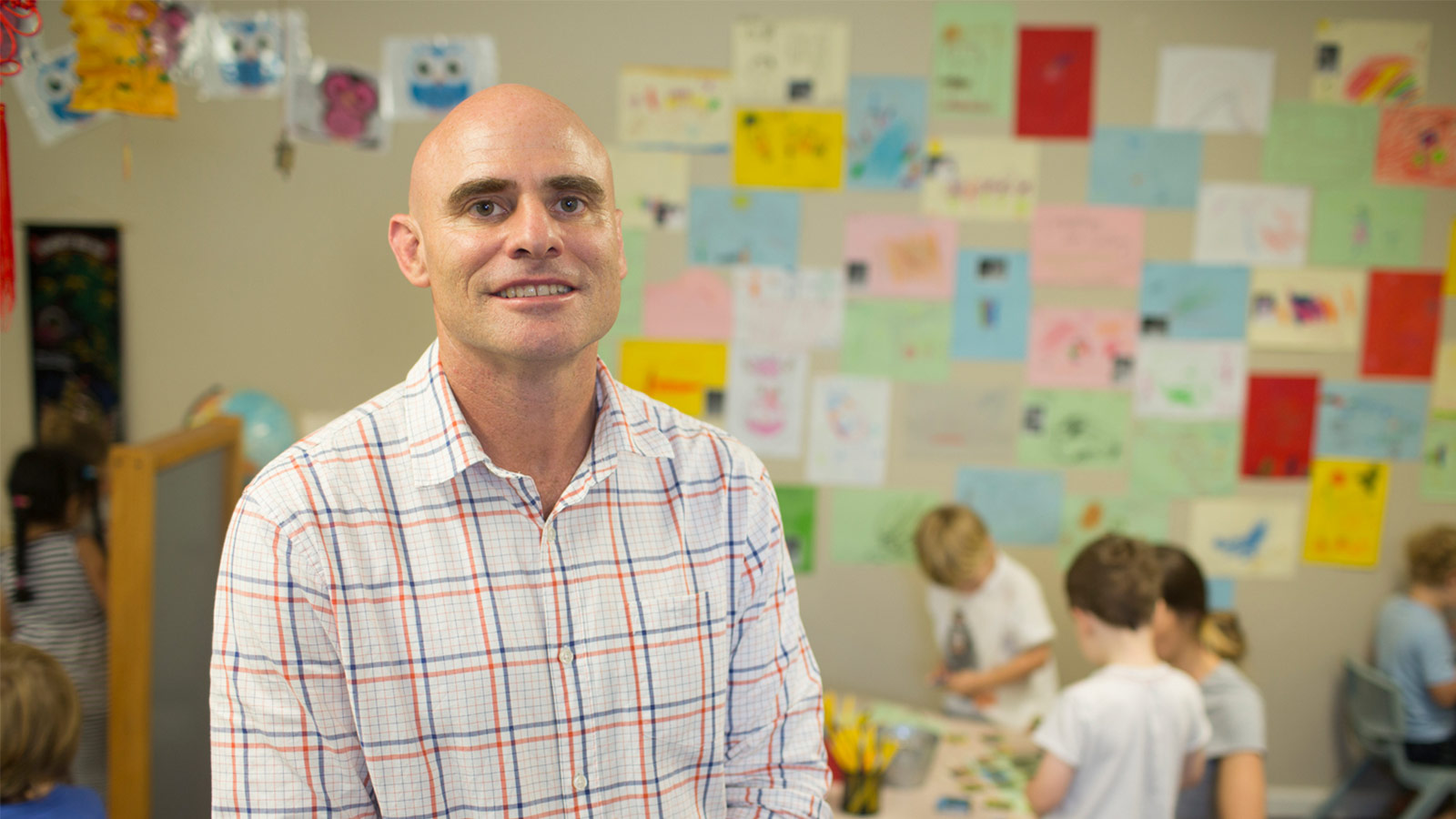 Professor Anthony Okely is wearing a red, blue and white tartan business shirt standing in a classroom. There are children doing deskwork behind him.