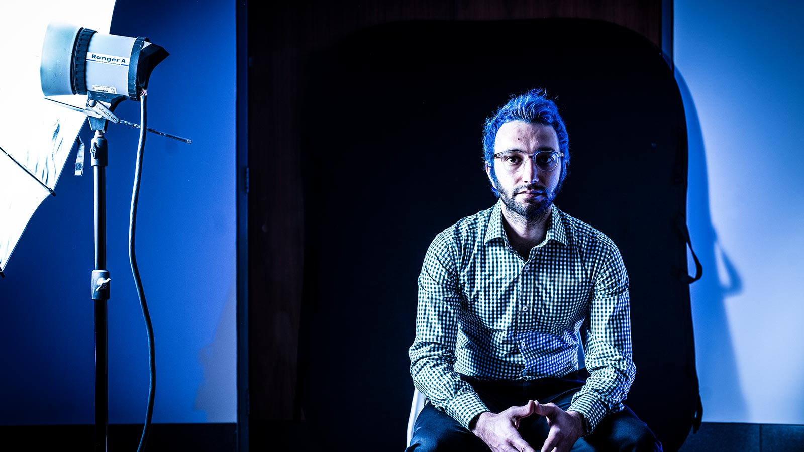 Dr Armin Alimardani has blue hair and clear glasses. He is wearing a checkered button-up shirt and sitting on a chair in a blue-lit studio. To his left is a photography flash light and umbrella.