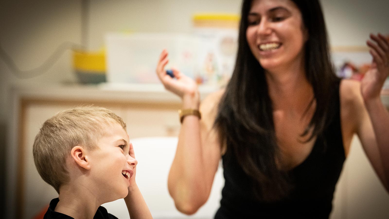 Ashleigh has tan skin and long dark hair. She is wearing a black top and playing with her young son Alex who has blond hair and is holding his hand up to his eye smiling.