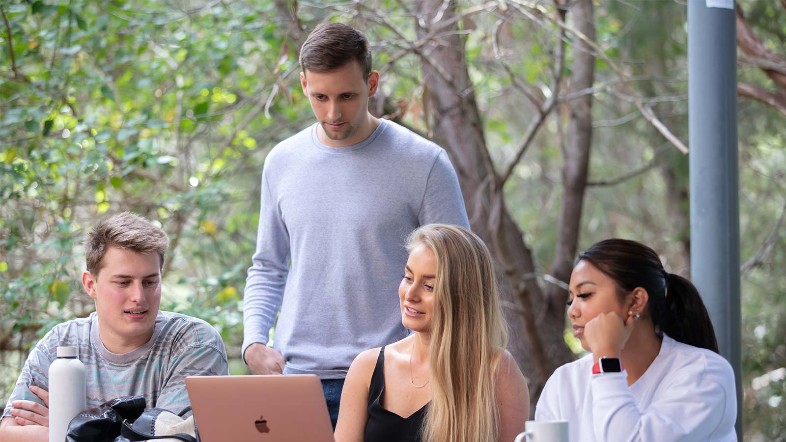 Three students are seated at an outdoor table looking at a laptop. There is a student standing behind them looking down at the laptop. There are green trees inn the background.