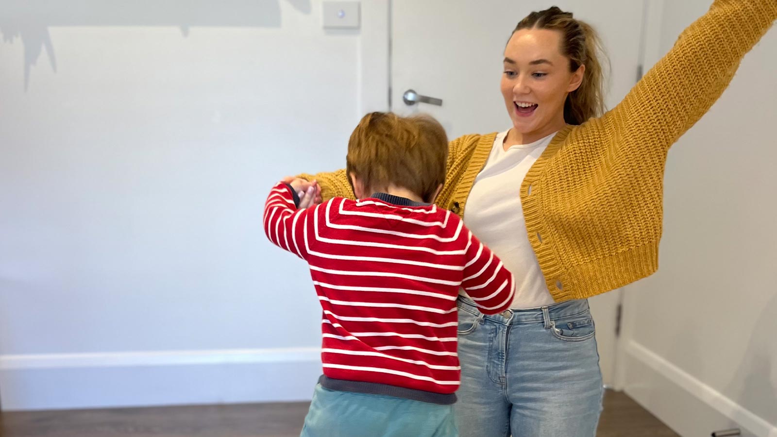 A white woman in a white t-shirt, blue jeans and yellow cardigan is playig with a toddler. She is on her knees with her arms in the air.