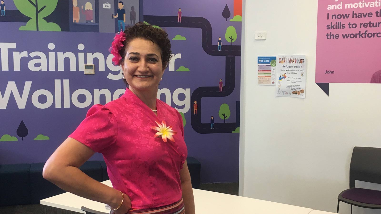 Didi Kello has tan skin and short dark hair. She is wearing a pink shirt and a pink flower in her hair, standing in a adult education classroom
