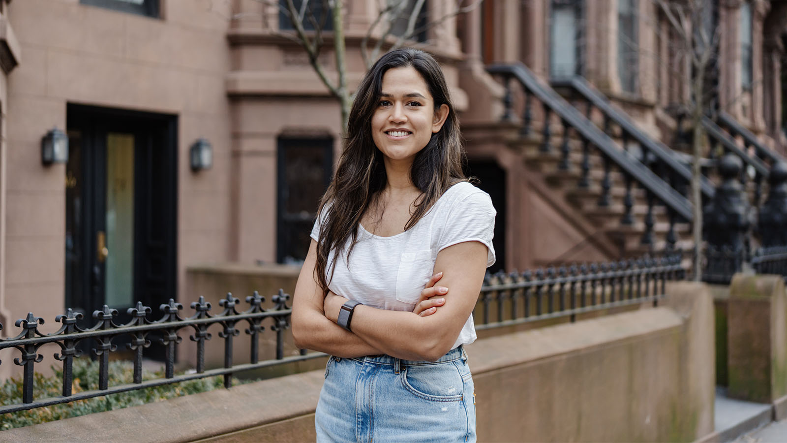 A woman with a white shirt and blue jeans is standing in front of a NYC brownstone building with her arms crossed, smiling