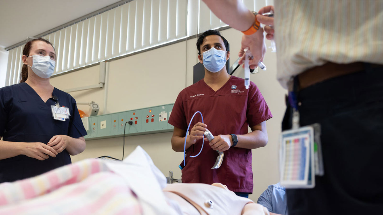 Two student doctors in maroon scrubs and PPE are being taught on a dummy patient in a simulation lab.