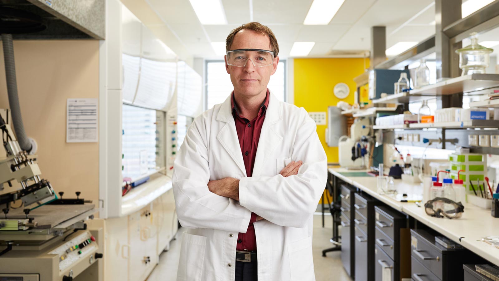 Geoff Spinks is standing in a lab, wearing a white labcoat and lab goggles with his arms crossed.