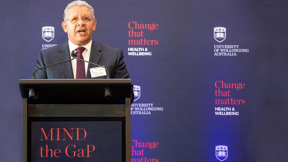 Glen Williams is standing at a podium in front of a UOW media wall. The podium has red text that reads MIND the GaP.
