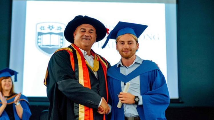Harry Clout has blond hair and a blond beard and is accepting his degree at graduation. He is wearing a blue gown and hat a and shaking hands with a man in a black gown and hat.