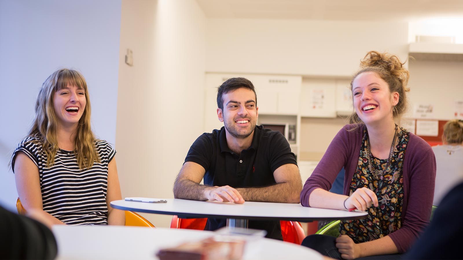 Three strudents sitting at a table in a communal area, smiling.