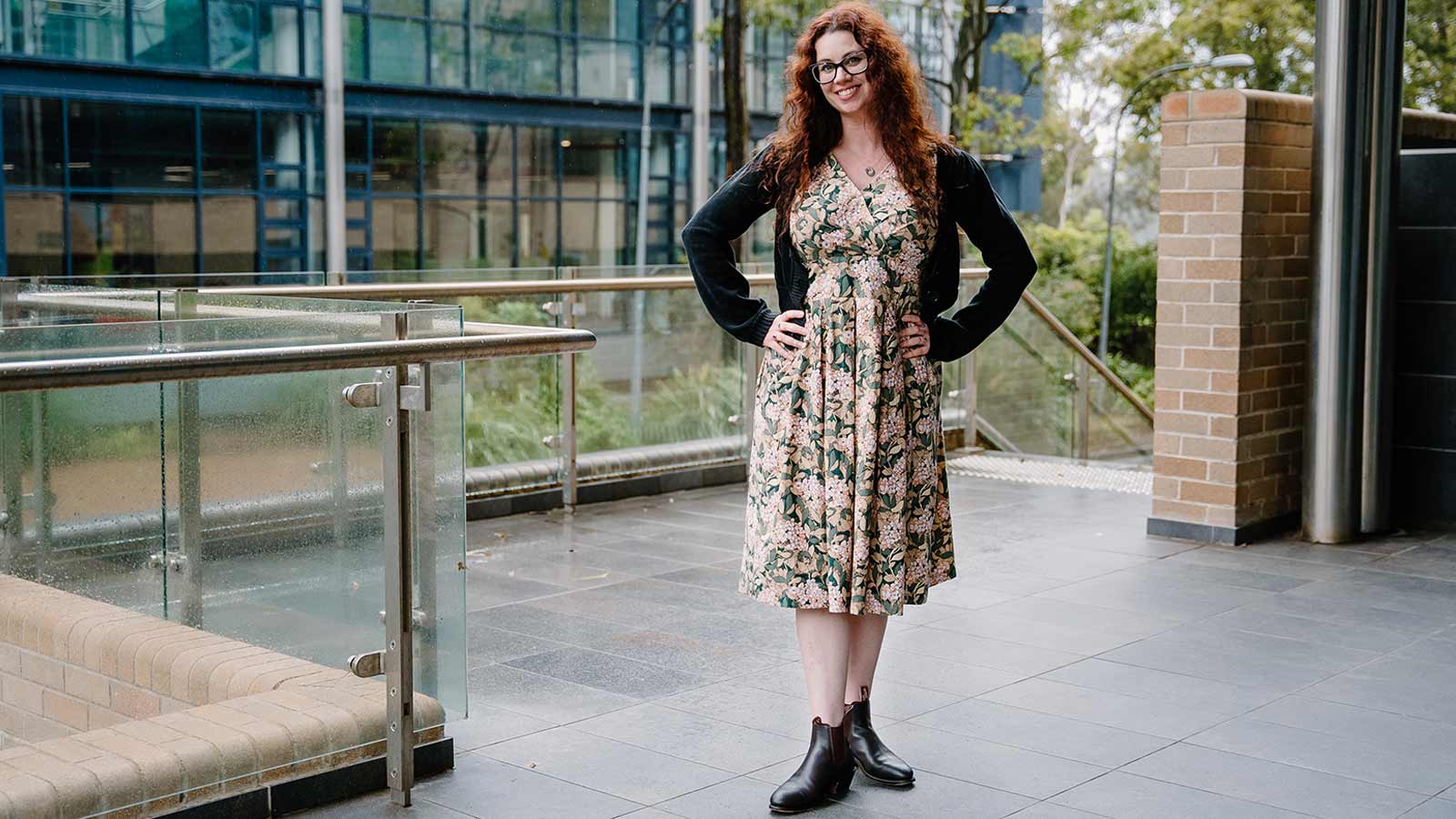 Molly Mackanzie is standing outside a building at UOW. She has pale skin and dark red hair and is wearing a floral dress, black cardigan and black boots.