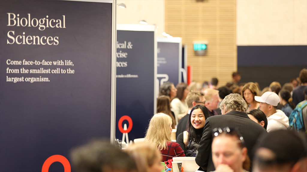 A crowd of students in a hall. There are navy blue UOW banners on the left, the front one reads 'Biological sciennces'.