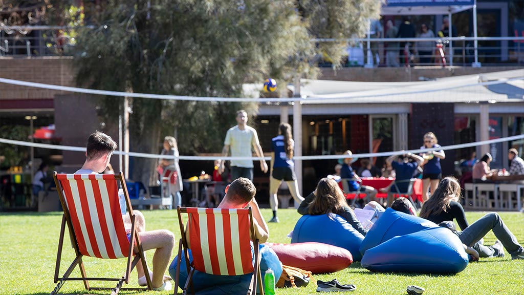 A group of students sitting on banana lounges and beanbags on McKinnon lawn. There are people playing badminton in the background.