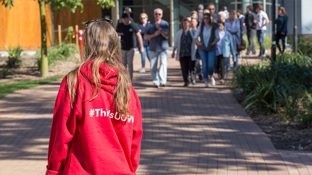 The back of a girl with long brown hair. She is wearing a red hoodie that says #ThisIsUOW, a crowd of people are walking towards her.