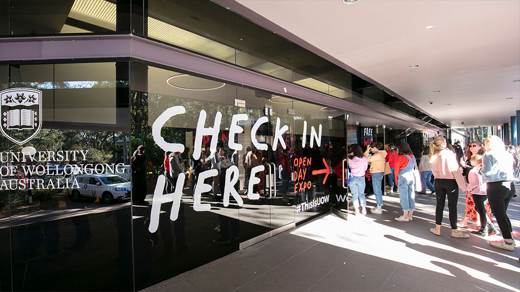 Side angle view of the UOW Uni Hall. A decal on the window reads 'Check in here' and students are walking in the door
