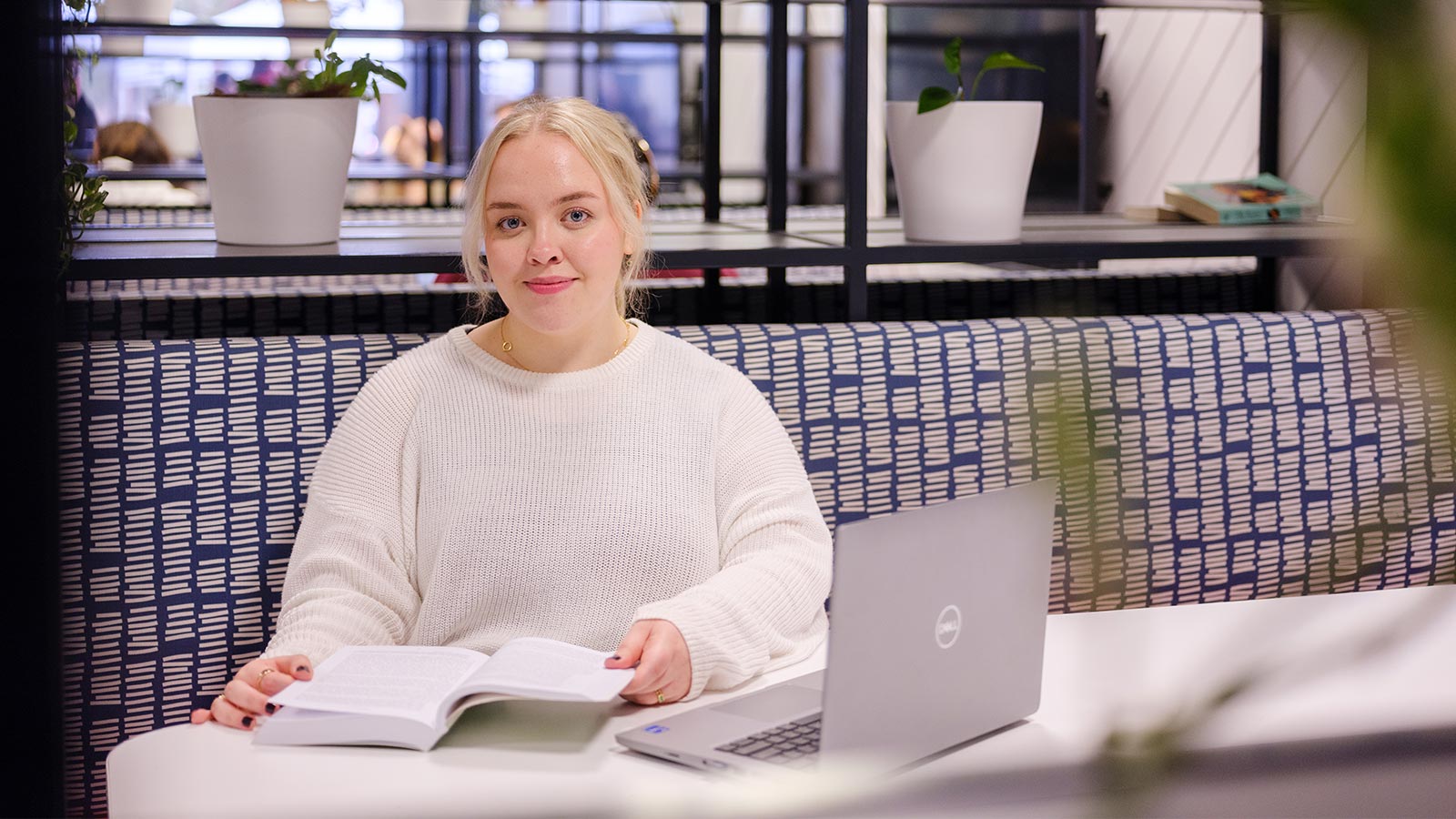 Siobhan Heywood has light tan skin and blond hair. She is sitting with a book and a Dell laptop at a white desk in the UOW library, surrounded by pot plants.