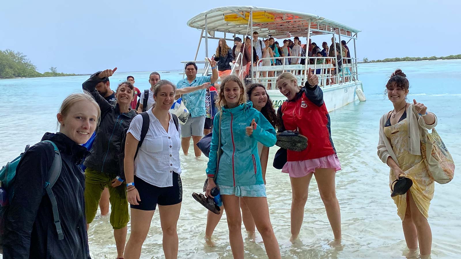 A group of students are standing in ankle deep ocean water in front of a charter boat in Tahiti.