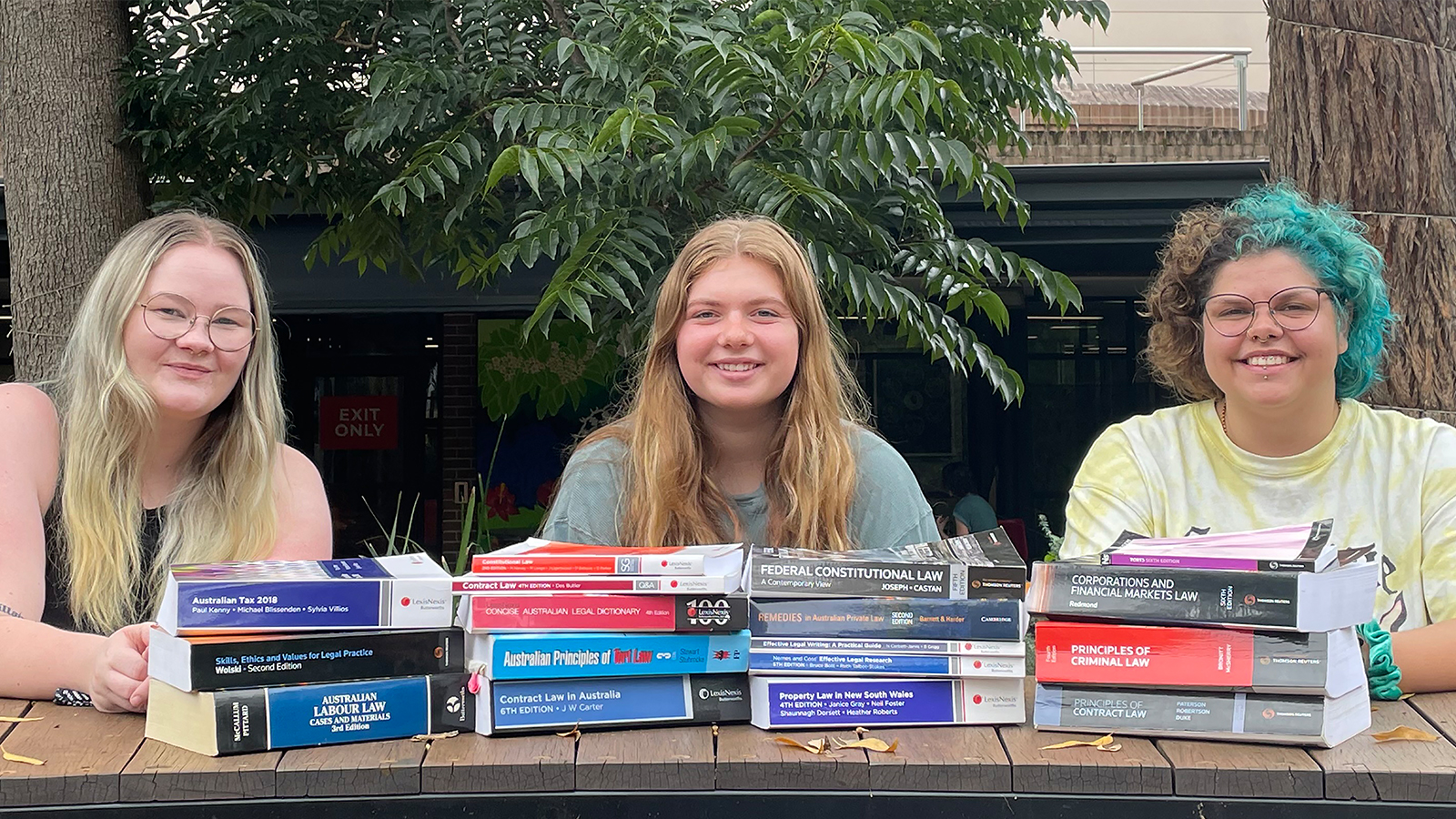 Three University of Wollongong Law students sit at an outdoor bench smiling, surrounded by law textbooks