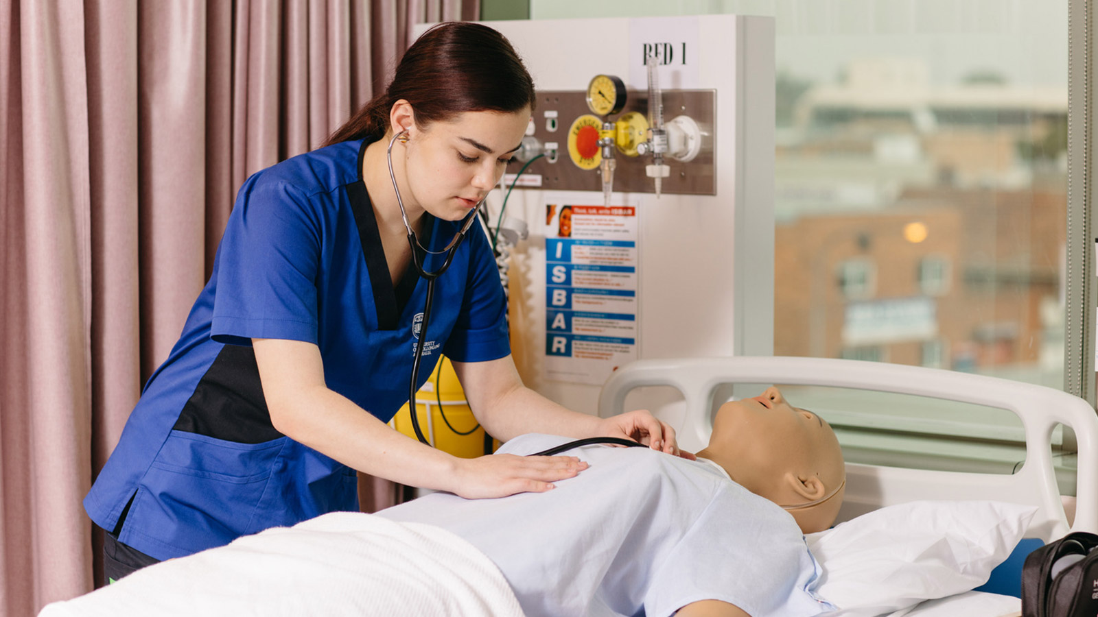 Angelique has pale skin and dark brown hair. She is in a nusing lab at UOW, wearing a dark blue uniform. She is using a stethoscope on a dummy patient.