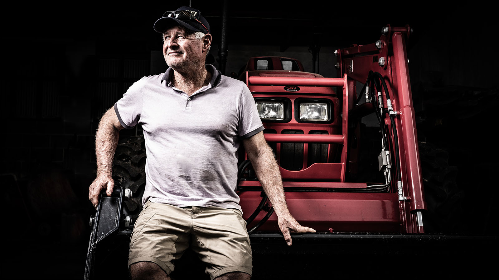 A man in a grey polo shirt and black cap is standing sitting against a red tractor