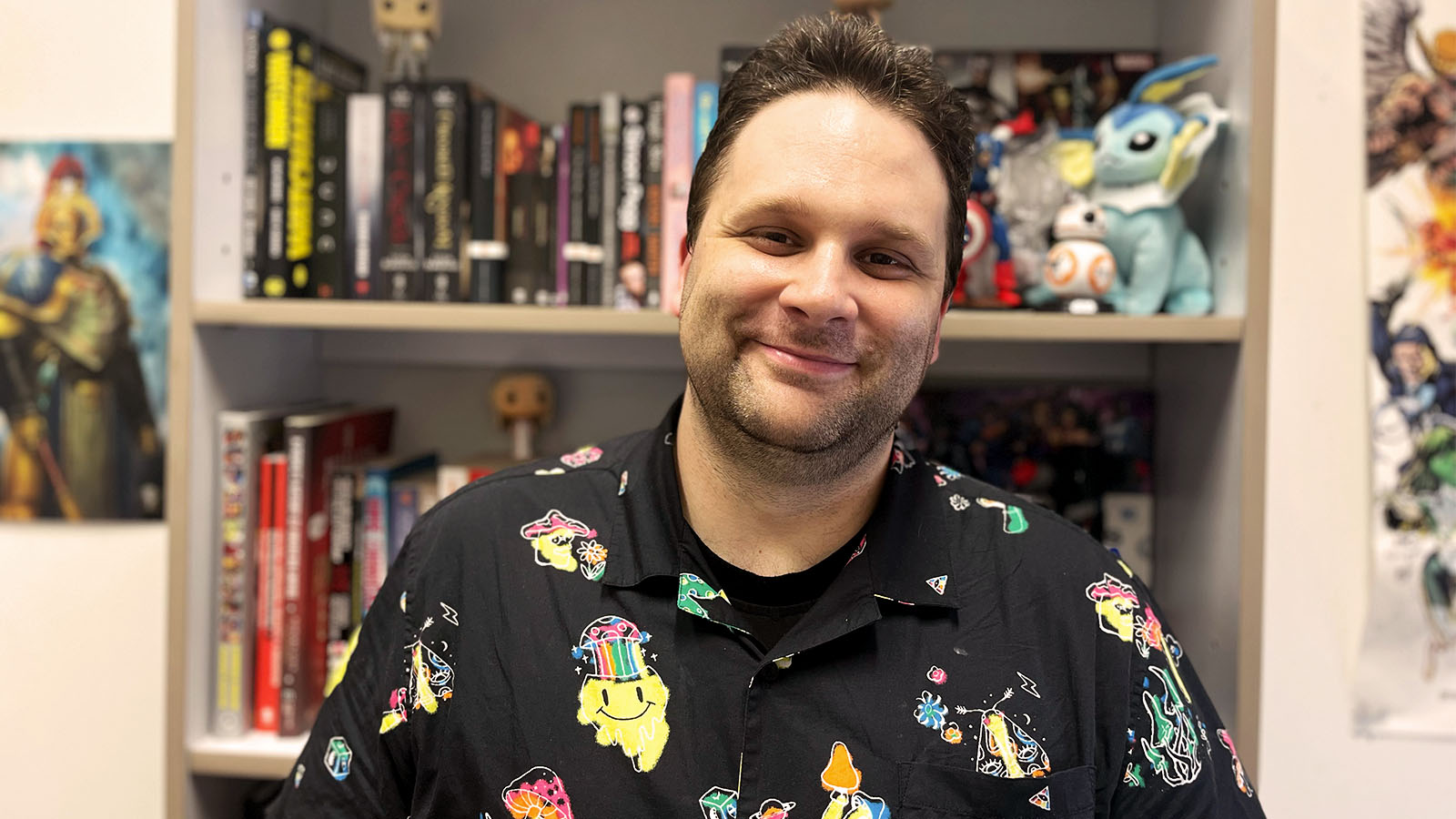 A man is wearing a black shirt with colourful cartoon drawings on it. He is tanding in front fo a bookshelf stacked with books and figurines.