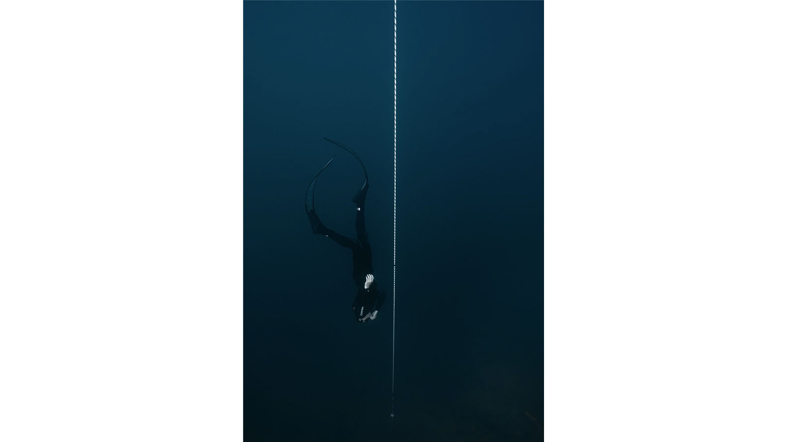 A man is free diving downwards in the ocean, parallel to a safety line