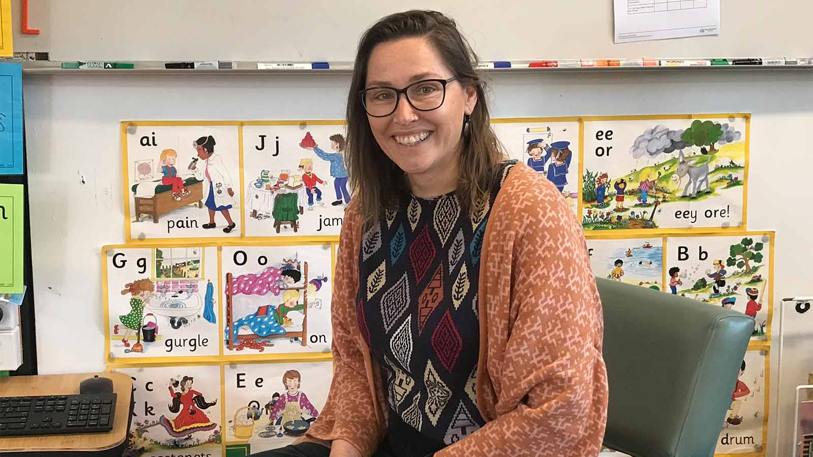 Danielle has brown hair and glasses. She is wearing a black and white top and orange cardigan, sitting on a small classroom chair in front of an alphabet chart.